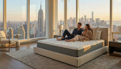 Two people sitting on a mattress in a room with large windows offering a cityscape view.